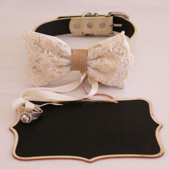 White lace ring bearer dog bow tie collar featuring floral embroidered lace, burlap center detail, attached wedding ring holder ribbon, and decorative ring next to a blank wedding signboard backdrop