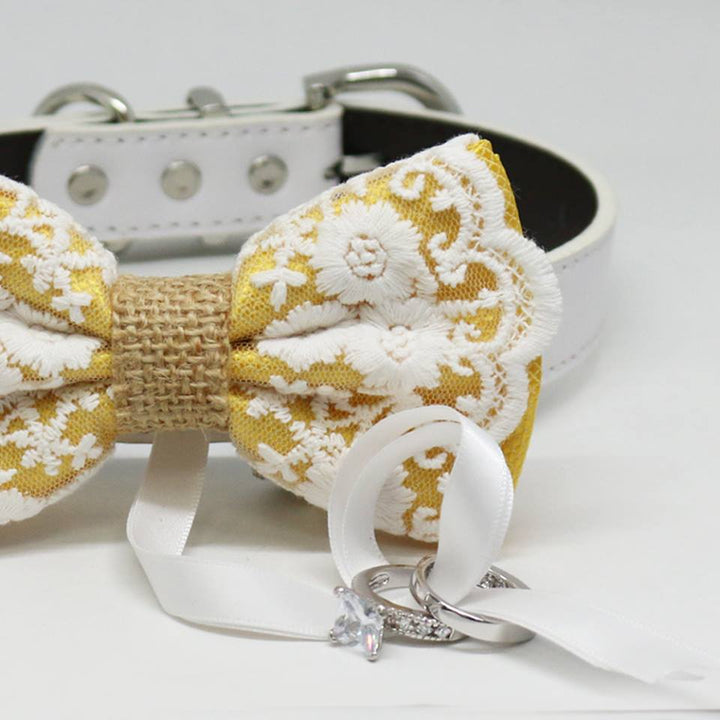 Close-up detail of yellow lace ring bearer dog bow tie with floral embroidered lace overlay, attached wedding ring holder ribbon, decorative ring set, and white vegan leather collar